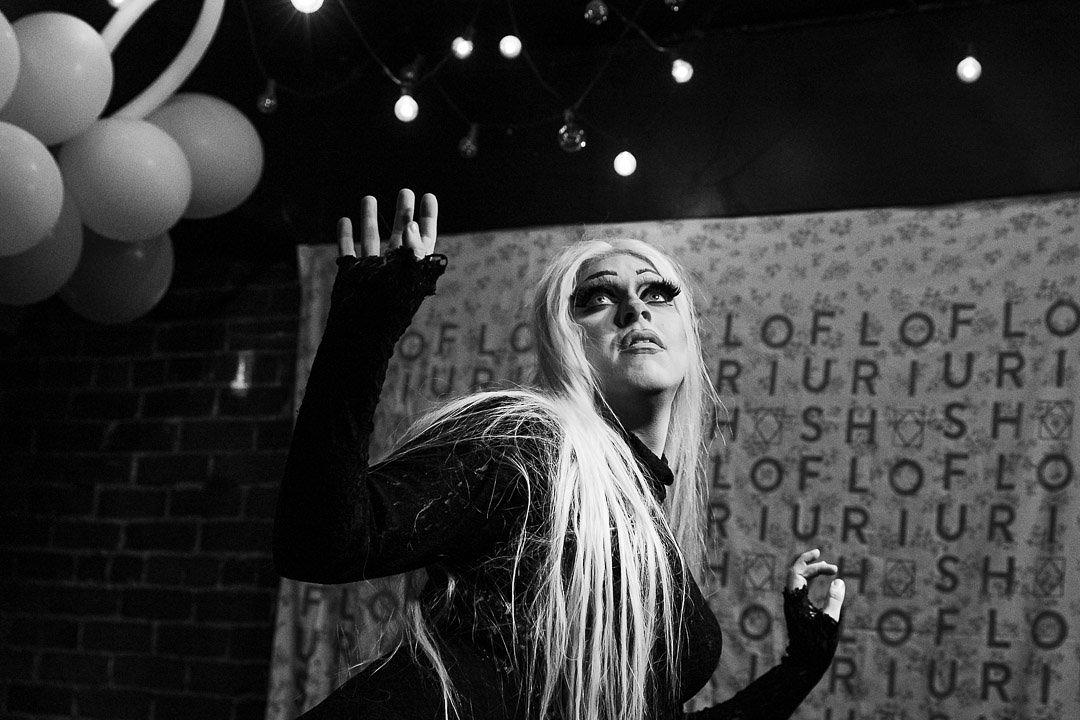 Black and white photo of drag artist Luna Lafleur on stage in front of a FLOURISH Festival backdrop and balloons - long blonde hair with dramatic black eye makeup. Luna looks longingly up at the ceiling with her hands outstretched. 